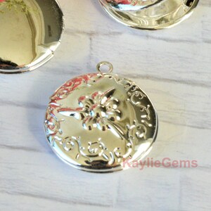 Op de afbeelding: Zilverkleurige ronde medaillon met een bloemmotief. De medaillon heeft een verhoogde bloem in het midden en decoratief krulwerk langs de rand. Bovenop zit een kleine lus voor een ketting. De tekst "KaylieGems" is zichtbaar. Diameter ongeveer 3 cm.