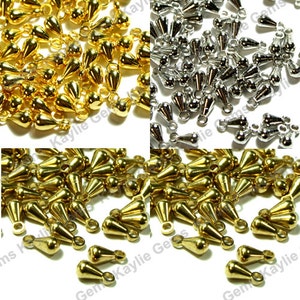 May include: A large quantity of gold and silver teardrop-shaped metal beads with a loop at the top. The beads are arranged in a random pattern on a white background.