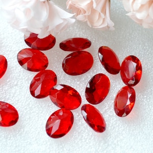 May include: Oval-shaped, faceted red gemstones scattered on a white surface. The gemstones are a deep, transparent red, reflecting light. Soft pink and white flowers are in the background. These could be used for jewelry making or other crafts.