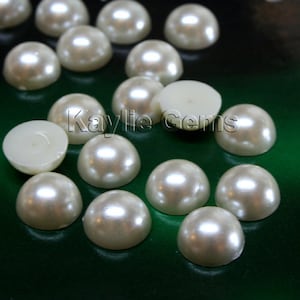 May include: A collection of off-white pearl cabochons, some with the flat back showing. The pearls are arranged on a green surface.