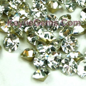 Puede incluir: Un montón grande de piedras de cristal redondas, transparentes y facetadas. Las piedras tienen un acabado plateado y están esparcidas sobre una superficie blanca.