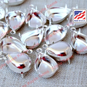 May include: A group of silver-toned metal teardrop-shaped settings with prongs on the bottom and a loop at the top. The settings are arranged on a textured fabric surface. Made in the U.S.A.