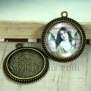 May include: Two round metal pendant trays with a decorative edge. One tray is empty and the other has a glass cabochon with an image of a young girl with angel wings.
