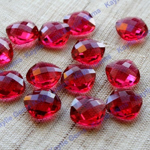May include: A group of 14 red faceted glass heart-shaped beads. The beads are scattered on a beige fabric surface.