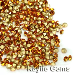 May include: A large pile of orange and gold rhinestones. The rhinestones are scattered on a white surface. The rhinestones are small and have a pointed back.
