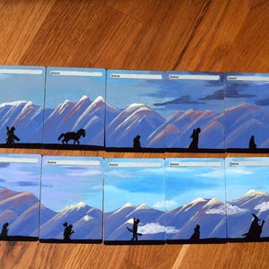 Può includere: Nove dipinti di paesaggi raffiguranti scene di montagna con figure e un cavallo in silhouette. Ogni dipinto ha un cielo blu e montagne innevate. La parola "Forest" è scritta nella parte superiore di ogni dipinto.