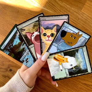 Könnte beinhalten: Eine Hand hält eine Sammlung von handbemalten Magic: The Gathering-Karten. Die Karten zeigen skurrile Tierporträts, darunter eine Katze, ein Hund und eine Ente, vor Landschaftshintergründen. Die Karten werden auf einer Holzoberfläche präsentiert.