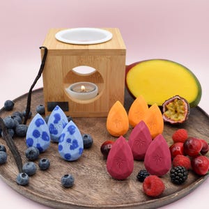 Könnte beinhalten: Ein Holz-Aromatherapie-Diffusor mit einem weißen Keramikdeckel und einer brennenden Kerze im Inneren. Der Diffusor ist von verschiedenen Früchten umgeben, darunter eine Mango, eine Passionsfrucht und Blaubeeren. Es gibt auch mehrere kleine, bunte Wachsschmelzen in Tropfenform mit Palmenmotiven.