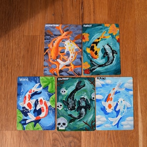 Könnte beinhalten: Fünf handgemalte Kunstkarten mit Koi-Karpfen in verschiedenen aquatischen Umgebungen. Jede Karte trägt den Namen einer Umgebung: Berg, Wald, Insel, Sumpf und Ebenen. Die Koi-Karpfen sind in Orange, Weiß, Schwarz und Blau gemalt.
