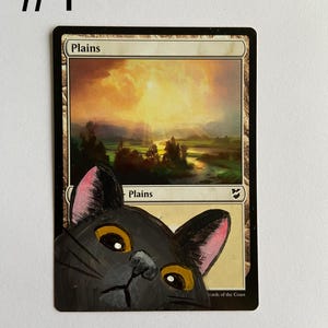 May include: A hand-painted black cat peeking over a "Plains" Magic: The Gathering card. The card features a landscape with a sunset. The cat has yellow eyes and pink inner ears. The number "#1" is in the upper left corner.