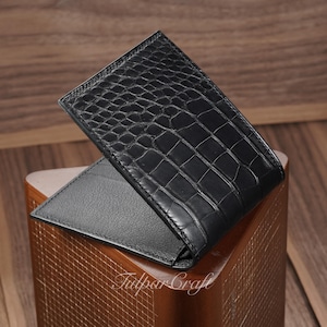 Könnte beinhalten: Eine schwarze Lederbrieftasche mit Krokodilhautstruktur. Die Brieftasche ist geöffnet und zeigt ein schwarzes Innenleben. Die Brieftasche wird auf einer Holzoberfläche präsentiert. Der Markenname "Tulpar Craft" ist sichtbar.