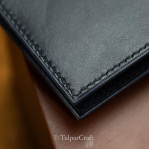 Op de afbeelding: Close-up van een zwarte leren portemonneehoek. Het leer heeft een gladde textuur met zichtbare stiksels langs de randen. De portemonnee lijkt goed gemaakt en toont een minimalistisch ontwerp.