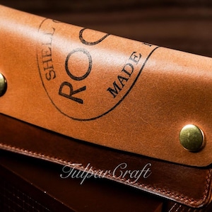 Op de afbeelding: Close-up van een handgemaakt lederen item in bruine tinten. Het item heeft de tekst "ROC MADE" in een cirkelvormige stempel. Messingkleurige metalen drukknopen bevestigen het item. De tekst "Tulpar Craft" is ook zichtbaar.