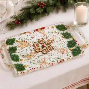 May include: A rectangular serving tray with clear handles, decorated with Christmas-themed elements. The tray features green holly leaves, gold stars, and small red berries. It also has gold illustrations of houses and reindeer, and small gingerbread cookies.