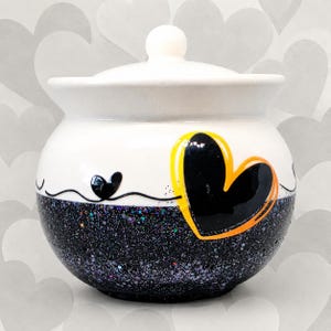 May include: A white ceramic jar with a lid, featuring a black glittery base and a hand-painted design. The design includes a large black heart outlined in yellow and orange, alongside a smaller black heart connected by a thin black line.
