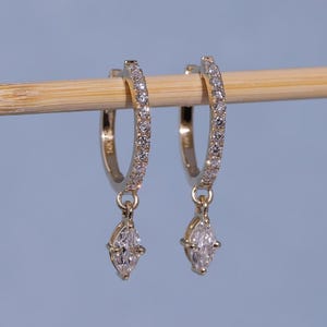 May include: A pair of gold hoop earrings with a row of small, clear stones along the hoop. Each earring has a teardrop-shaped, clear stone pendant. The earrings are displayed on a light brown wooden dowel against a light blue background.