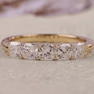 May include: A gold band ring featuring five clear, square-cut gemstones. The ring is set on a neutral surface with a blurred background. The ring's design is simple, elegant, and suitable for various occasions.