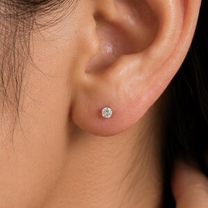 May include: A small, round, clear stud earring featuring a single faceted gemstone set in a metallic prong setting, positioned centrally on the earlobe. The earring has a brilliant, reflective surface that catches the light, creating a sparkle.