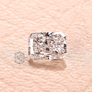 May include: A radiant-cut diamond with a rectangular shape, showcasing internal facets and brilliance. The diamond is set against a soft, textured, pinkish-beige background. A small logo is visible in the lower left corner.
