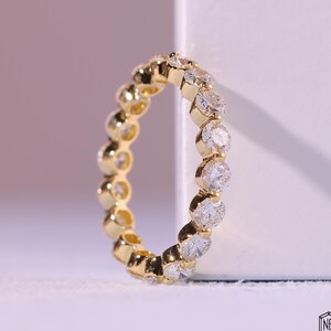 Op de afbeelding: Een gouden ring met een doorlopende rij ronde, heldere edelstenen. De band van de ring is ook goudkleurig, met een gestructureerd, afgerond ontwerp tussen elke edelsteen. De ring staat tegen een zachte, vage achtergrond.