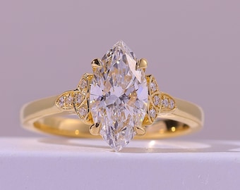 Puede incluir: Un anillo de compromiso de oro con un diamante talla marquesa. El anillo presenta un diamante grande y transparente como pieza central, con diamantes más pequeños engastados a los lados. La banda es de color oro pulido y el anillo está sobre un fondo suave y neutro.