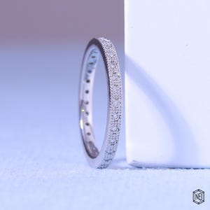 May include: A silver-toned eternity band ring featuring a continuous row of small, round, clear stones set along the outer edge. The metal band has a textured, milgrain-style border detail framing the stones, creating a refined and classic appearance.