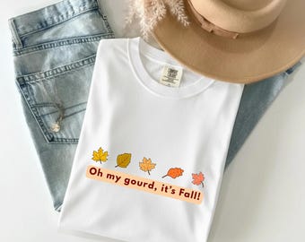 May include: White t-shirt with the text "Oh my gourd, it's Fall!" and colorful leaf graphics. The shirt is paired with light-wash jeans, a tan hat, and a necklace.