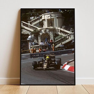May include: A black and gold Formula One race car with the words "John Player Special" on the side, driving on a track with a white line and red and white curbing. The car is in the foreground, and a building with a staircase is in the background.