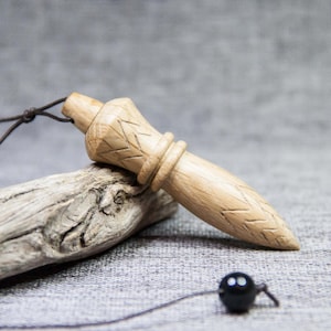 Peut inclure: Un pendule en bois clair avec des détails sculptés et une perle noire au bout d'une ficelle. Le pendule repose sur un morceau de bois flotté sur un fond gris.