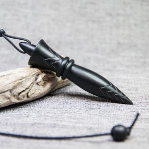 May include: A black wooden pendulum with a pointed tip and intricate carvings. The pendulum is attached to a black cord with a small black bead at the end.