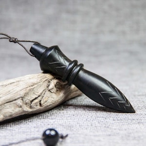 Peut inclure: Pendentif en bois noir sculpté, avec une extrémité pointue et des gravures décoratives en forme de feuilles. Le pendentif est suspendu à un cordon fin et brun foncé. Il repose sur un morceau de bois flotté.