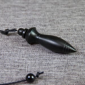 May include: A black wooden pendulum with a pointed tip and a string attached. The pendulum is on a gray fabric surface.