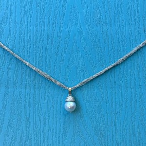 Può includere: Una collana a catena d'argento con un ciondolo a perla singola. La perla è bianca e ha una leggera lucentezza iridescente. La catena è composta da più fili di filo d'argento.