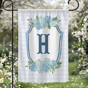 May include: A garden flag with a light blue and white gingham pattern. The flag features a white shield with the letter "H" in dark blue, surrounded by blue hydrangea flowers and green leaves. The flag is displayed on a black metal stand.