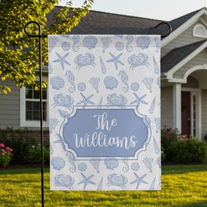 May include: A personalized garden flag featuring a light blue and white seashell pattern. The flag has a light blue oval with the name "The Williams" in white script. The flag is displayed in a front yard.