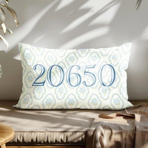 May include: A rectangular cushion featuring a floral pattern in light blue and green on a cream backdrop. The number "20650" is displayed in a large, elegant blue font across the centre. The cushion rests on a neutral-toned surface, with shadows cast across it.