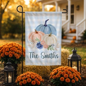 Puede incluir: Una bandera de jardín rectangular con un patrón de cuadros vichy en azul claro y blanco. El diseño muestra tres calabazas en azul, melocotón y blanco, acentuadas con flores azules y melocotón y hojas verdes. El texto The Smiths está en negro.