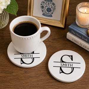 May include: Two round white ceramic coasters featuring a black printed design of a large letter S bisected by a horizontal line with the name SMITH centered inside. One coaster supports a white ceramic mug filled with black coffee.