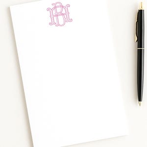 Könnte beinhalten: Ein weißer, rechteckiger Notizblock mit einem rosafarbenen Monogramm der Buchstaben B und H in der oberen Mitte. Ein schwarzer Kugelschreiber mit goldfarbenen Akzenten befindet sich rechts neben dem Notizblock. Das Papier ist unter dem Monogramm leer.