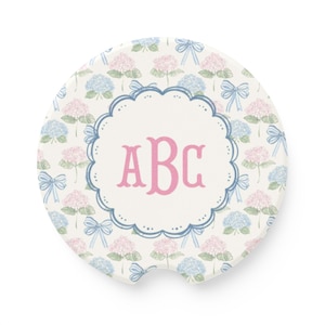 May include: A circular car coaster featuring a pattern of pink and blue hydrangea flowers with light blue bows on a white background. The center has a scalloped blue frame enclosing the pink letters ABC in a serif font with a notch at the bottom.