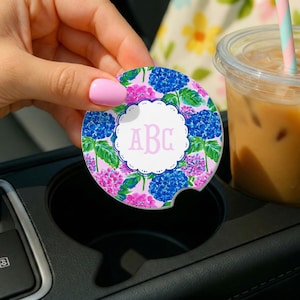 May include: A round car cup holder coaster featuring a vibrant floral pattern of blue and pink hydrangea blossoms with green leaves. The center displays a white scalloped circular frame containing the light pink monogram letters ABC.