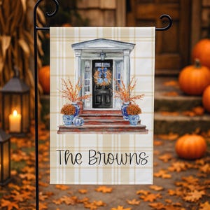 May include: A rectangular garden flag featuring a light yellow and white plaid background. The center displays a watercolor illustration of a front porch with a black door, a wreath, and blue and white planters. The text The Browns is printed below.