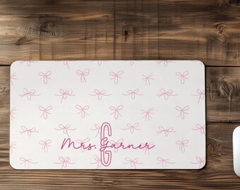 May include: A rectangular desk mat featuring a white background with a repeating pattern of light pink hand-drawn bows. The center of the mat displays the text Mrs. Garner in a dark pink script font, accompanied by a large, outlined letter G.