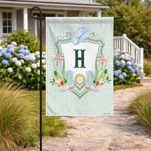 Puede incluir: Una bandera de jardín rectangular con un patrón de cuadros vichy en verde claro y blanco. El centro presenta un escudo blanco con una letra H en verde oscuro, rodeado de ilustraciones en acuarela de un sombrero vaquero y botas azules.