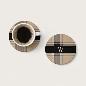 May include: Two round coasters featuring a beige, black, and thin red plaid pattern. One coaster supports a white ceramic mug filled with black coffee. The other coaster displays a centred, capitalised white letter W on a horizontal black stripe.