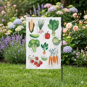 Può includere: Una bandiera da giardino bianca con illustrazioni ad acquerello di varie verdure, tra cui mais, aglio, spinaci, un peperone rosso, broccoli, carote e pomodori. La bandiera è esposta all'aperto in un giardino.