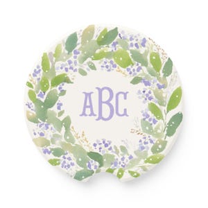 May include: A round ceramic car coaster featuring a watercolor wreath of green leaves and small purple flowers. The center of the coaster displays the letters ABC in a light purple serif font. The coaster has a small semi-circular notch at the bottom.