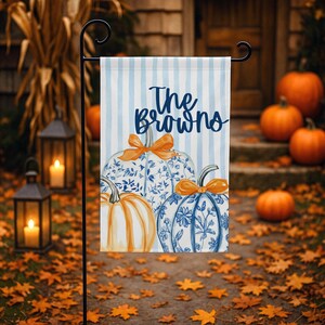 May include: A rectangular garden flag featuring a light blue and white vertical striped background. The text The Browns is printed in dark blue script. Below the text are three pumpkins, two with blue and white floral patterns and one solid orange.