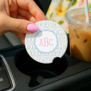 May include: A circular car coaster featuring a light blue and green damask-style floral pattern with small pink flowers. The center has a white scalloped circle containing the pink monogram letters ABC in a serif font, with a notched edge.