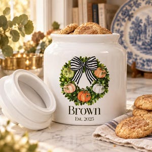 Peut inclure: Un pot à biscuits en céramique blanche avec couvercle, orné d'une couronne de feuilles vertes et de citrouilles orange, blanches et vertes, surmontée d'un nœud rayé noir et blanc. Le texte indique Brown Est. 2025 en police serif.